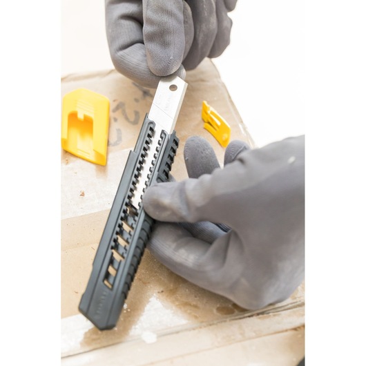 Photo of STANLEY 0-10-151 snap-off knife with black handle and gray blade, held by gloved hands, with yellow covers nearby.