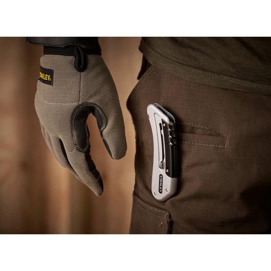 STANLEY 0-10-810 folding utility knife clipped to brown pants pocket, close-up side view, with gloved hand nearby.