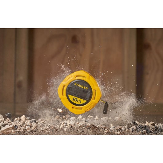 Stanley 0-34-295 tape measure, yellow casing, 10m, shown on rocky ground with dust in the air.