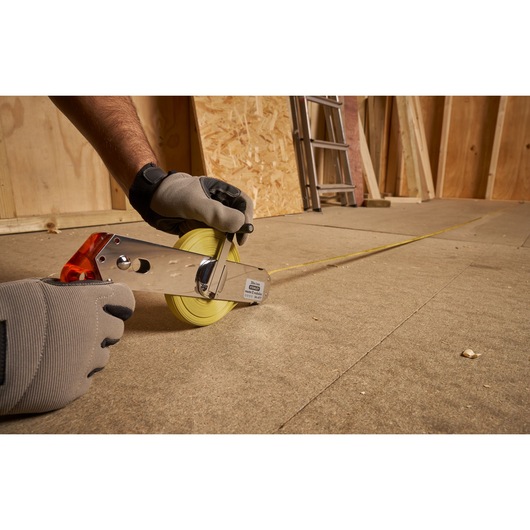 STANLEY 1-34-477 open reel tape measure with yellow blade, used by gloved hands on a construction floor.