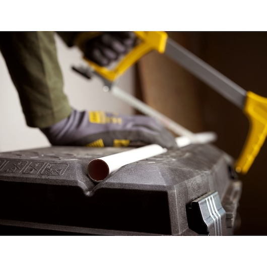 STANLEY 1-94-210 toolbox lid used as a work surface, cutting white plastic pipe with yellow hacksaw, close-up.