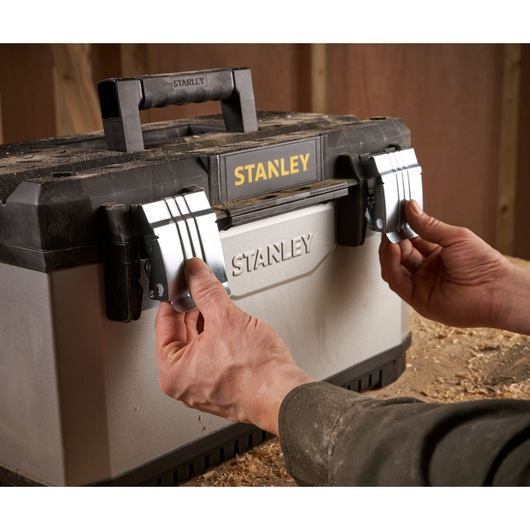 Photo of STANLEY 1-95-615 toolbox with hands opening metal latches, view from front in workshop setting.