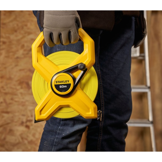 STANLEY 2-34-795 measuring tape 60m held by person wearing gloves, close-up, indoor work setting.