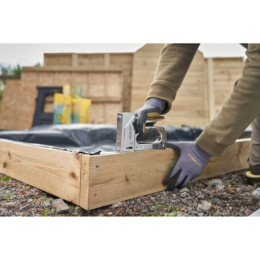 Photo of STANLEY 6-TR110 stapler being used to fasten plastic liner to wooden garden bed frame outdoors.