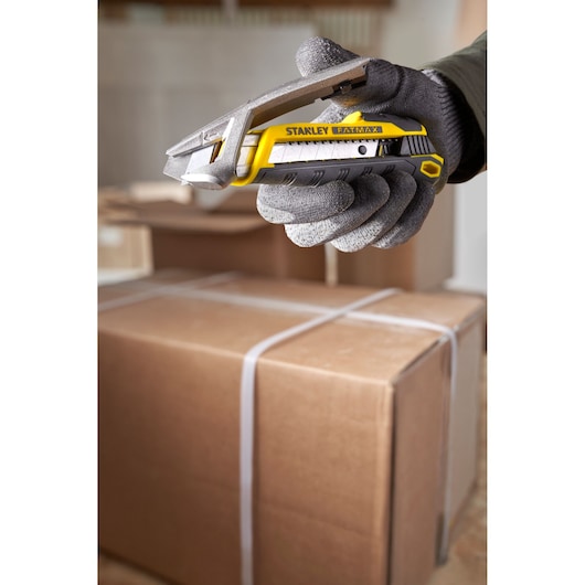 Stanley FMHT10594-0 utility knife held in gloved hand, opening a cardboard box in warehouse.