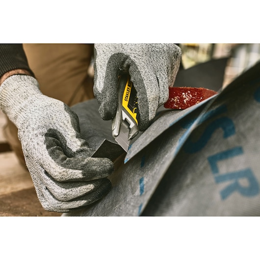 Photo of STANLEY FMHT10594-0 utility knife cutting fabric, close-up with gloved hands.