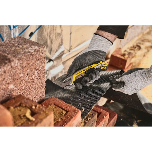 STANLEY FMHT10594-0 utility knife cutting black material near bricks, close-up photo.