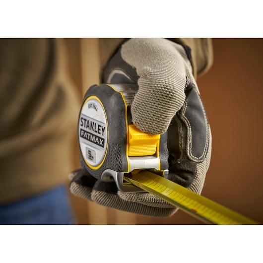 Photo of STANLEY FMHT38214-0 FatMax tape measure held by gloved hand, yellow and black body, extended blade.