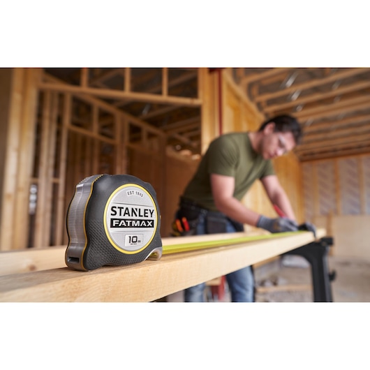 STANLEY FATMAX 10m tape measure FMHT38232-0 on wood beam, construction worker measuring in background.