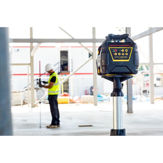 STANLEY FMHT77449-1 rotary laser level on tripod at construction site, worker in background.