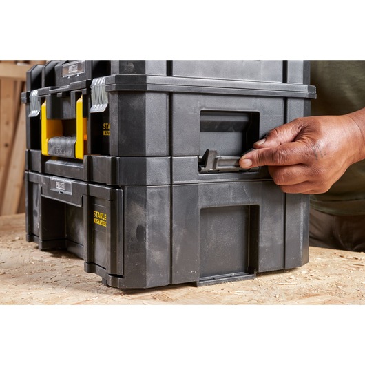 STANLEY FMST1-71968 black stackable toolbox with yellow handles, photography showing latch being opened by hand.