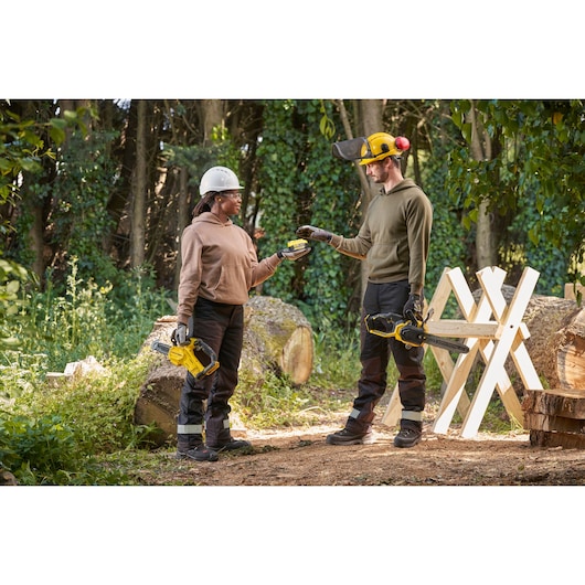 STANLEY SFMCPS415D1 cordless chainsaw in use outdoors, two people exchanging battery, logs in background.