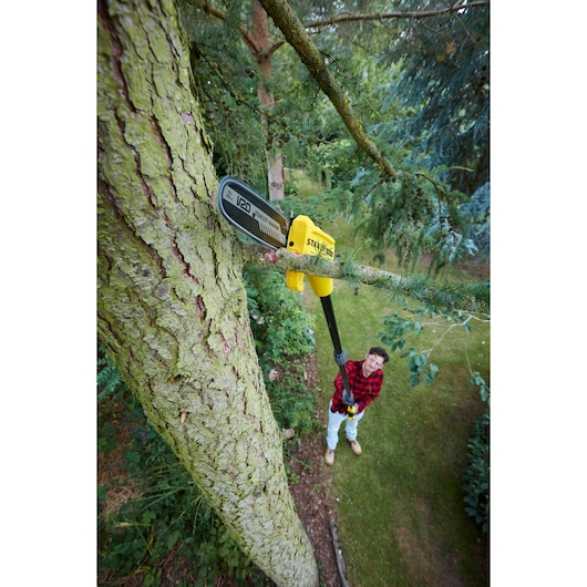 STANLEY SFMCPS620M1 pole saw cutting high tree branch in garden, user in plaid shirt operating from below.