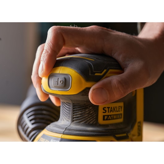 Photo of STANLEY SFMCW220B sander, close-up of hand pressing power button, yellow and black design.