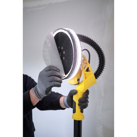 STANLEY SFMEE500S drywall sander close-up with gloved hands changing sanding pad, white background.