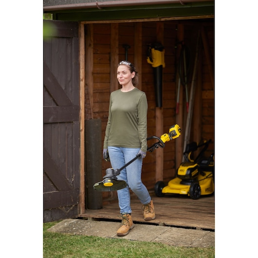 STANLEY SFMSTB930B string trimmer held by a person in workwear, standing outside a wooden shed with garden tools.