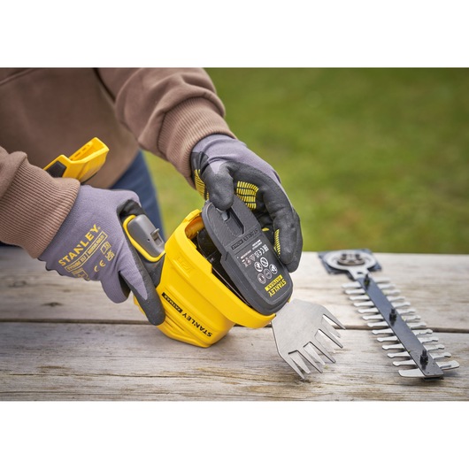 STANLEY STCSS20D1 cordless shrub and grass shear being fitted with battery, gloves worn, parts on wooden table.