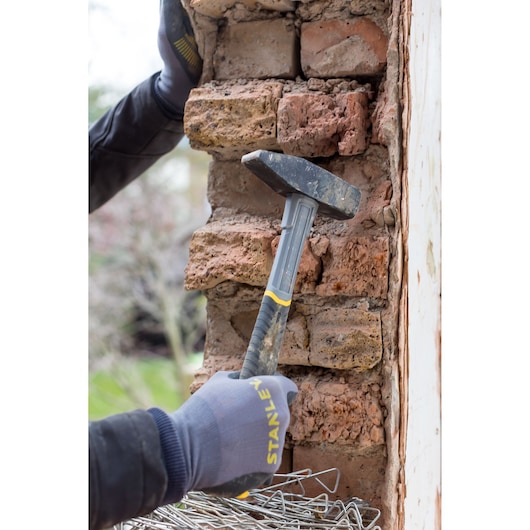 STANLEY STHT0-51909 club hammer held by gloved hand, used for masonry demolition on a brick wall, side view.
