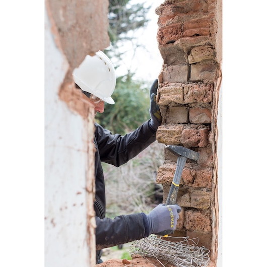 Photo of a worker using a STANLEY STHT0-51909 hammer on a brick wall, wearing gloves and a hard hat outdoors.