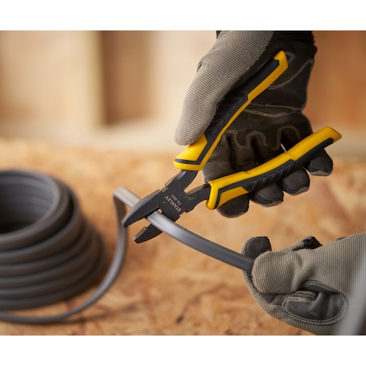 STANLEY STHT0-74454 pliers with yellow and black handle cutting thick gray wire, close-up, worn by gloved hands.