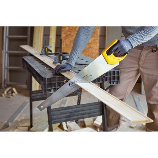 STANLEY STHT1-20353 hand saw with yellow handle cutting wood plank in workshop setting.