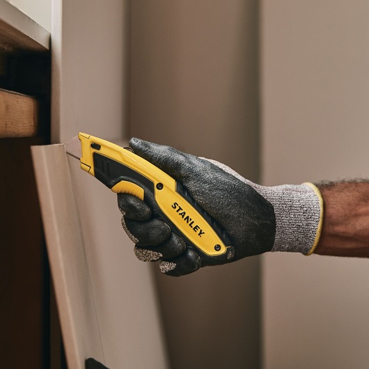 Stanley STHT10479-0 utility knife held in gloved hand, cutting a panel. Photography product category.