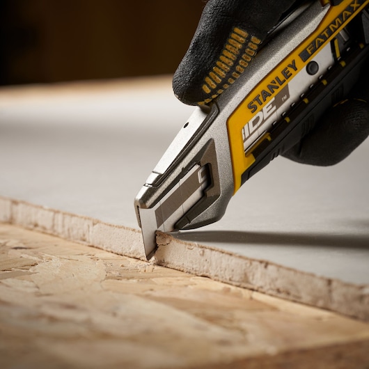 Photo of STANLEY STHT2-11818 utility knife cutting drywall, close-up on blade and hand in glove.