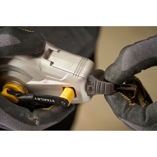 Close-up photo of STANLEY STHT47611-0 tape measure, metallic body, yellow lever, gloved hands operating clip.