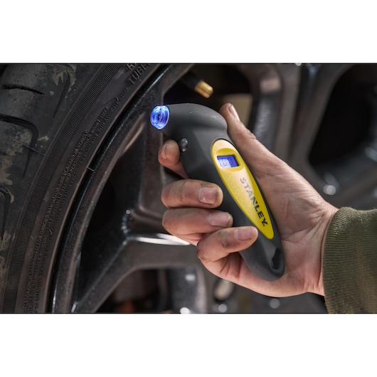 STANLEY STHT80874-0 digital tire pressure gauge checking tire, close-up of hand holding tool with blue display.