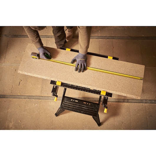 STANLEY STST83400-1 workbench with worker measuring wood plank using tape measure in workshop.